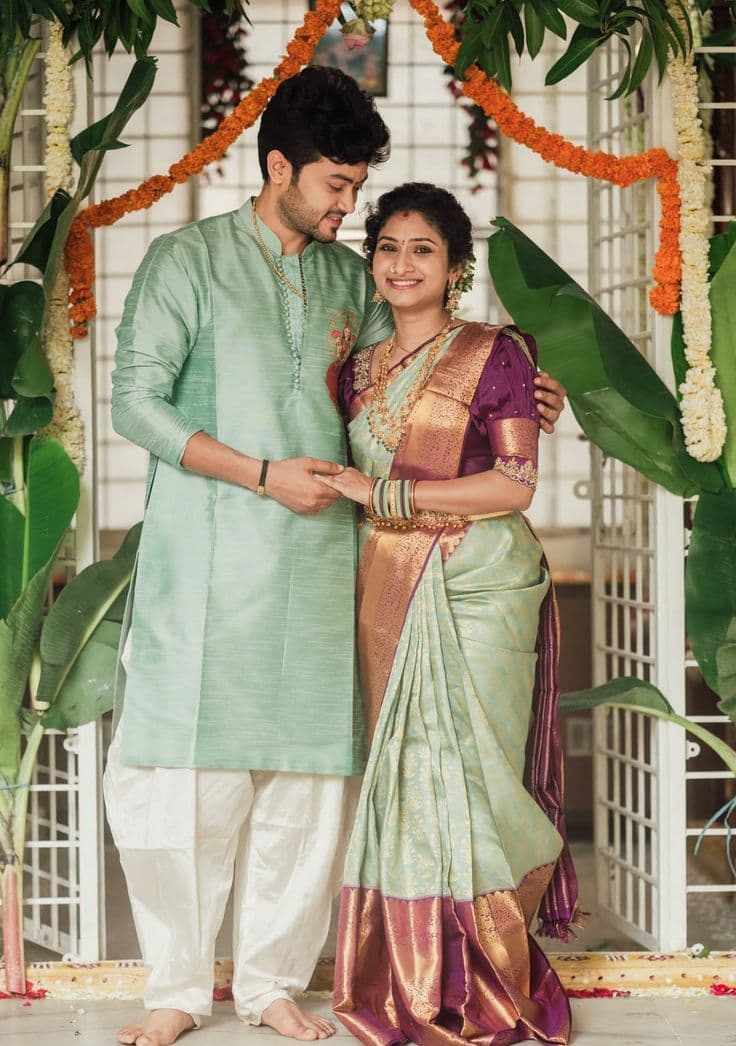 Smiling couple in silk wedding attire beneath a garland of marigolds